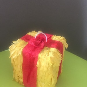 May include: A yellow, fringed, cube-shaped piñata with a red ribbon wrapped around it. A white plastic loop is attached to the top. The piñata is sitting on a green surface.