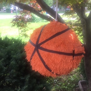 Puede incluir: Una piñata de baloncesto naranja con rayas negras cuelga de una rama de árbol. La piñata está hecha de papel con flecos y está suspendida por una cuerda blanca. El fondo incluye follaje verde y un vistazo a una zona residencial.