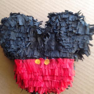 May include: A handmade piñata in the shape of a cartoon character's head. The ears are black, and the face is red with two yellow circles. The piñata is made of fringed paper.