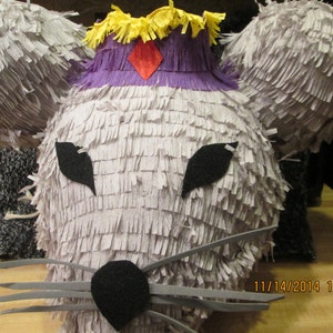 Puede incluir: Una piñata gris y esponjosa con forma de ratón con una corona morada y amarilla. La piñata tiene ojos de fieltro negro y una nariz de fieltro negro. La piñata es una divertida decoración de fiesta.
