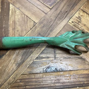 May include: A vintage, green metal garden tool with a wooden handle. The tool has a claw-like head with multiple tines and a long, slender handle. The green paint shows signs of wear, giving it a rustic appearance.