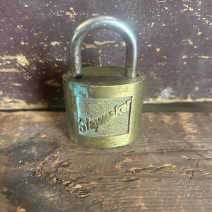 May include: A vintage brass padlock with a silver-colored shackle. The word "Slaymaker" is embossed on the front. The lock shows signs of age and wear, with a slightly tarnished appearance. The padlock is a classic security item.