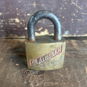May include: Vintage brass padlock with a dark metal shackle. The padlock has the word "SLAYMAKER" printed in red on a white background. The lock is sitting on a weathered wooden surface.