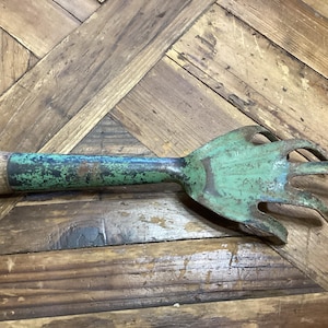 May include: An antique garden hand rake with a wooden handle and a green, claw-like head. The metal head shows signs of age and wear, with a weathered green finish. The wooden handle is light brown.
