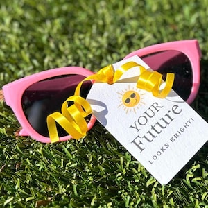 May include: Pink sunglasses with dark lenses rest on green grass, accompanied by a tag with the text "YOUR FUTURE LOOKS BRIGHT!" and a yellow ribbon. The tag features a smiling sun graphic.