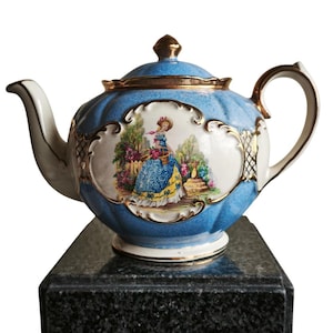 Puede incluir: Una tetera vintage de porcelana azul y blanca con detalles dorados. La tetera presenta una ilustración detallada de una mujer en un jardín. El asa y el pico son blancos, y la tapa está rematada con un adorno dorado.