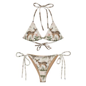 May include: A two-piece bikini set featuring a cream-colored background with a woodland print. The halter-style top and the bottoms are adorned with a pattern of deer, flowers, and ferns. The bikini has tie closures.