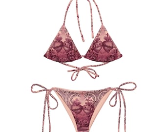 May include: A two-piece bikini set with a unique pattern. The top is a halter style, and the bottoms are tie-side. The fabric features a repeating design of hearts, crosses, and floral elements in shades of pink and burgundy.