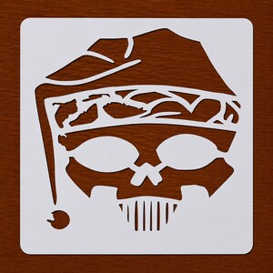 May include: A white stencil featuring a skull wearing a jester's hat. The skull has large eye sockets and a vertical line of teeth. The stencil is on a dark brown wooden surface.