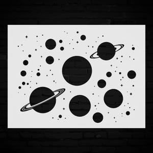 May include: A white stencil featuring a black space-themed design. The design includes planets, some with rings, and various sizes of stars and celestial bodies. The stencil is rectangular.