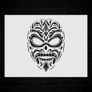 May include: Black and white tribal mask design. The mask features a symmetrical pattern with large eye shapes, a prominent nose, and a detailed mouth. The design is set against a white background, with a dark brick wall in the background.