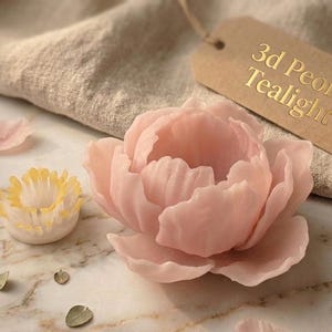 Puede incluir: Una vela de té de peonía 3D de color rosa claro con delicados pétalos. También hay una vela más pequeña con centro amarillo y pétalos dispersos. La etiqueta dice "3d Peony Tealight". Las velas descansan sobre una superficie de mármol.