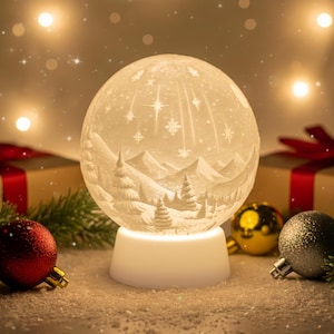 Könnte beinhalten: Weißer, kugelförmiger Weihnachtsschmuck mit einer detaillierten Winterlandschaft, einschließlich Berge und Bäume, die von einem sanften Schein beleuchtet wird. Der Schmuck steht auf einem weißen Sockel, umgeben von festlichen Dekorationen wie roten und goldenen Kugeln und Geschenkboxen.