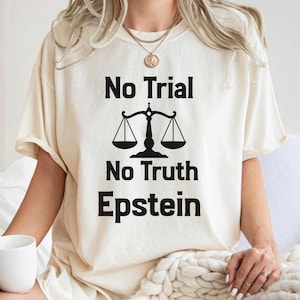 May include: Cream-colored t-shirt with the text "No Trial No Truth Epstein" in black, along with a scale graphic. The shirt is a relaxed fit, with short sleeves. The person is holding a white mug.