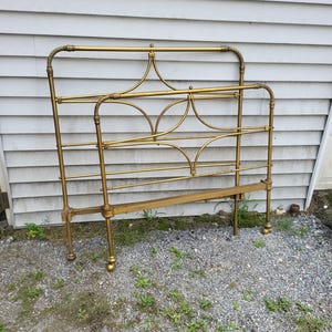 Op de afbeelding: Een vintage messing bedframe met een sierlijk ontwerp. Het hoofdbord en voetbord hebben gebogen lijnen en decoratieve accenten. Het frame is gemaakt van messing en heeft een warme, gouden kleur. Het bedframe is ontworpen voor een standaard matras.