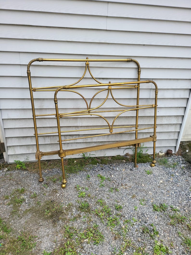 Op de afbeelding: Een vintage messing bedframe met een sierlijk ontwerp. Het hoofdbord en voetbord hebben gebogen lijnen en decoratieve accenten. Het frame is gemaakt van messing en heeft een warme, gouden kleur. Het bedframe is ontworpen voor een standaard matras.
