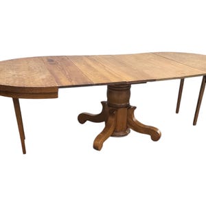 May include: An oval-shaped wooden dining table with a light brown finish. The table has a pedestal base with four curved feet and a tabletop that extends with leaves. The table is supported by four angled legs.