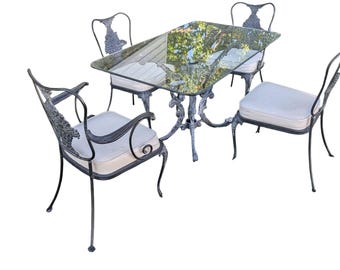 May include: A rectangular dining set featuring a glass-topped table with a dark metal frame and four matching chairs. Each chair has a dark metal frame with ornate scrollwork on the backrest and a light-colored, square seat cushion.