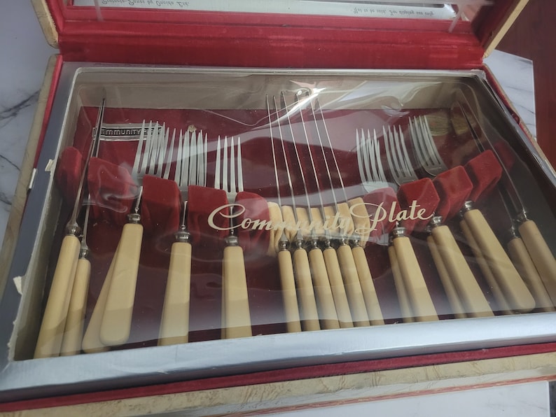May include: A wooden box with a red velvet interior and a clear plastic lid. The box contains a set of Community Plate silverware, including forks, knives, and spoons. The silverware is arranged in rows and is visible through the clear plastic lid.