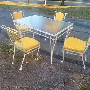 May include: White metal patio furniture set with a glass top table and four chairs with yellow cushions.