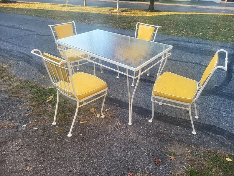 May include: White metal patio furniture set with a glass top table and four chairs with yellow cushions.