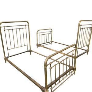 May include: A set of two antique-style brass bed frames featuring vertical spindle headboards and footboards with rounded top rails. The metal frames have a weathered, muted gold finish and include side rails for structural support.
