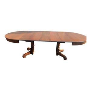 May include: A large, oval wooden dining table with a warm brown finish. The table features a split top with a leaf insert and ornate, carved pedestal legs. The table is approximately 72 inches long and 48 inches wide.