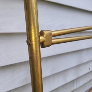 Op de afbeelding: Close-up van een goudkleurig metalen object met een cilindrische verticale steun en horizontale staven. Het metaal heeft een licht verouderd uiterlijk. Het object staat tegen een witte achtergrond met horizontale lamellen.