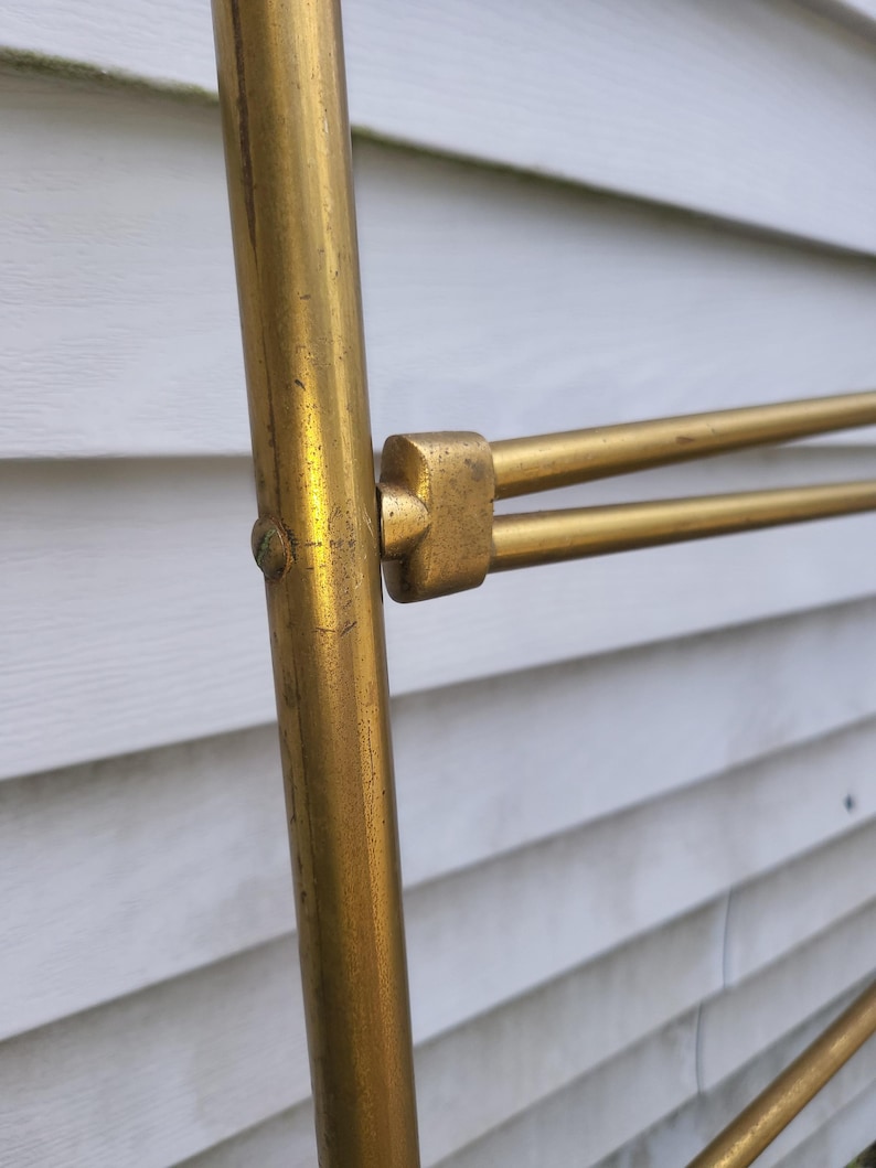 Op de afbeelding: Close-up van een goudkleurig metalen object met een cilindrische verticale steun en horizontale staven. Het metaal heeft een licht verouderd uiterlijk. Het object staat tegen een witte achtergrond met horizontale lamellen.