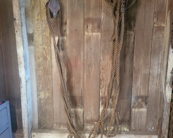 Puede incluir: Dos poleas de metal oxidadas con cuerda colgando de ellas, fijadas a una pared de madera. La cuerda es marrón y está enrollada.