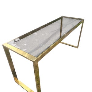 May include: A rectangular console table with a clear glass top and a gold-colored metal frame. The frame has a geometric design with angled legs. The table is designed for indoor use and is suitable for a living room or entryway.