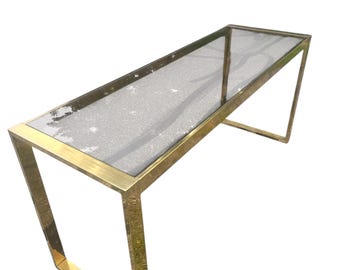 May include: A rectangular console table with a clear glass top and a gold-colored metal frame. The frame has a geometric design with angled legs. The table is designed for indoor use and is suitable for a living room or entryway.