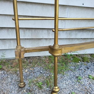 Op de afbeelding: Een vintage messing bedframe met een gouden afwerking. Het frame heeft verticale palen en horizontale staven, met decoratieve bolvormige voeten. Het bedframe is ontworpen voor een standaard matrasmaat.