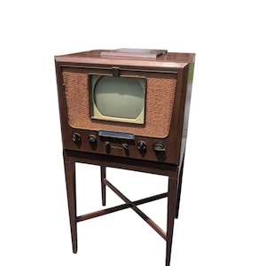 May include: A vintage television set with a dark brown wooden cabinet and a textured tan fabric speaker grille surrounding the screen. The unit features five black control knobs, a rectangular display, and stands on a tall, slender wooden base.