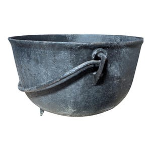 May include: A black cast iron cauldron with a rounded body, three small feet, and a curved handle attached to the rim. The cauldron has a textured surface and is designed for cooking or decorative purposes.