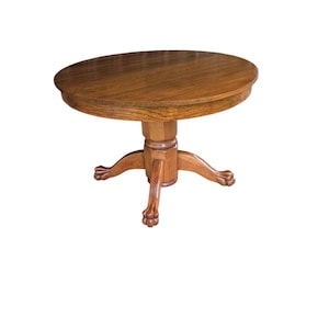 May include: A round wooden table with a medium brown finish and a visible wood grain pattern. The table features a central pedestal base that branches into four legs, each ending in a carved claw-and-ball foot design on a circular base.