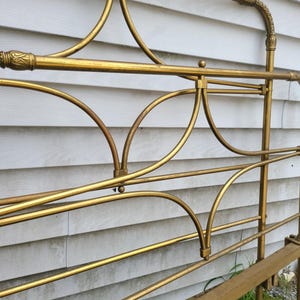 Op de afbeelding: Een close-up van een goudkleurig metalen bedframe. Het frame heeft gebogen en rechte lijnen, met decoratieve details aan de boven- en zijkanten. Het bedframe staat tegen een witte gevelbekleding.