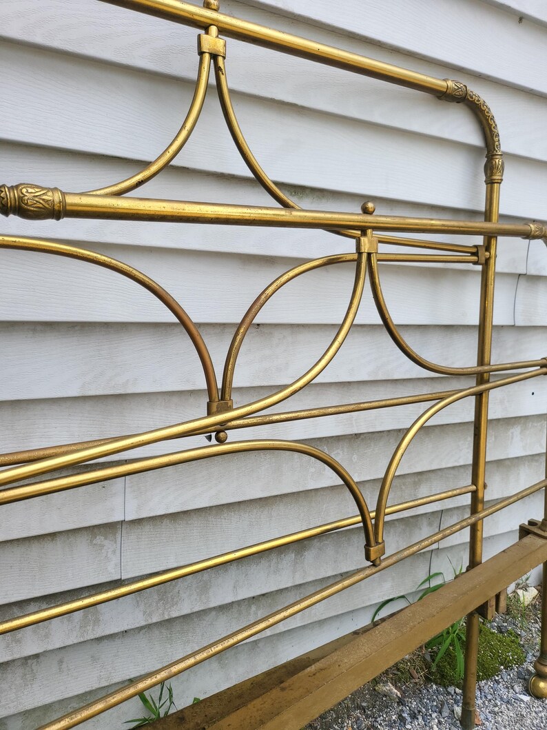 Op de afbeelding: Een close-up van een goudkleurig metalen bedframe. Het frame heeft gebogen en rechte lijnen, met decoratieve details aan de boven- en zijkanten. Het bedframe staat tegen een witte gevelbekleding.