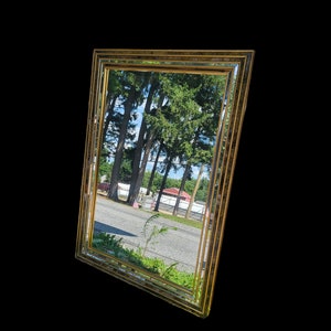 May include: A large, ornate gold-framed mirror with a beveled edge. The mirror reflects a view of trees and a road.