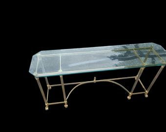 May include: A rectangular glass-top console table with a brass-colored metal frame. The table has an octagonal shape and features arched supports and decorative feet. The glass top is clear, and the frame has a polished finish.