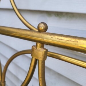 Op de afbeelding: Close-up van een gedeelte van een goudkleurig metalen bedframe. Het frame heeft een afgerond ontwerp met een kleine bal aan de bovenkant van een horizontale balk. Het metaal heeft een licht verouderd uiterlijk.