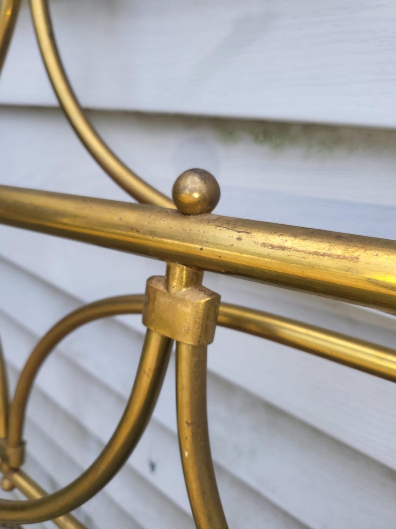 Op de afbeelding: Close-up van een gedeelte van een goudkleurig metalen bedframe. Het frame heeft een afgerond ontwerp met een kleine bal aan de bovenkant van een horizontale balk. Het metaal heeft een licht verouderd uiterlijk.