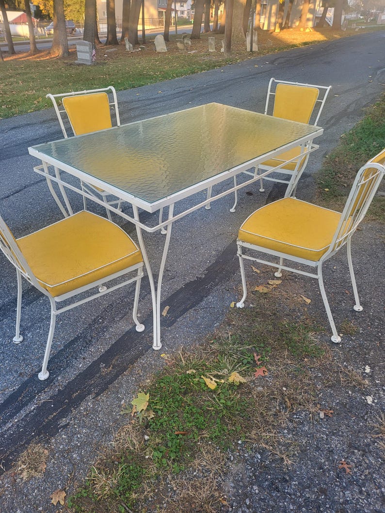 May include: A white metal patio dining set with four chairs and a glass-top table. The chairs have yellow cushions.