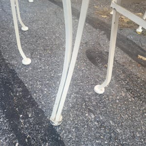 May include: Close-up of the white metal legs of a patio chair. The legs are curved and have a decorative design. The chair is sitting on a gray asphalt surface.