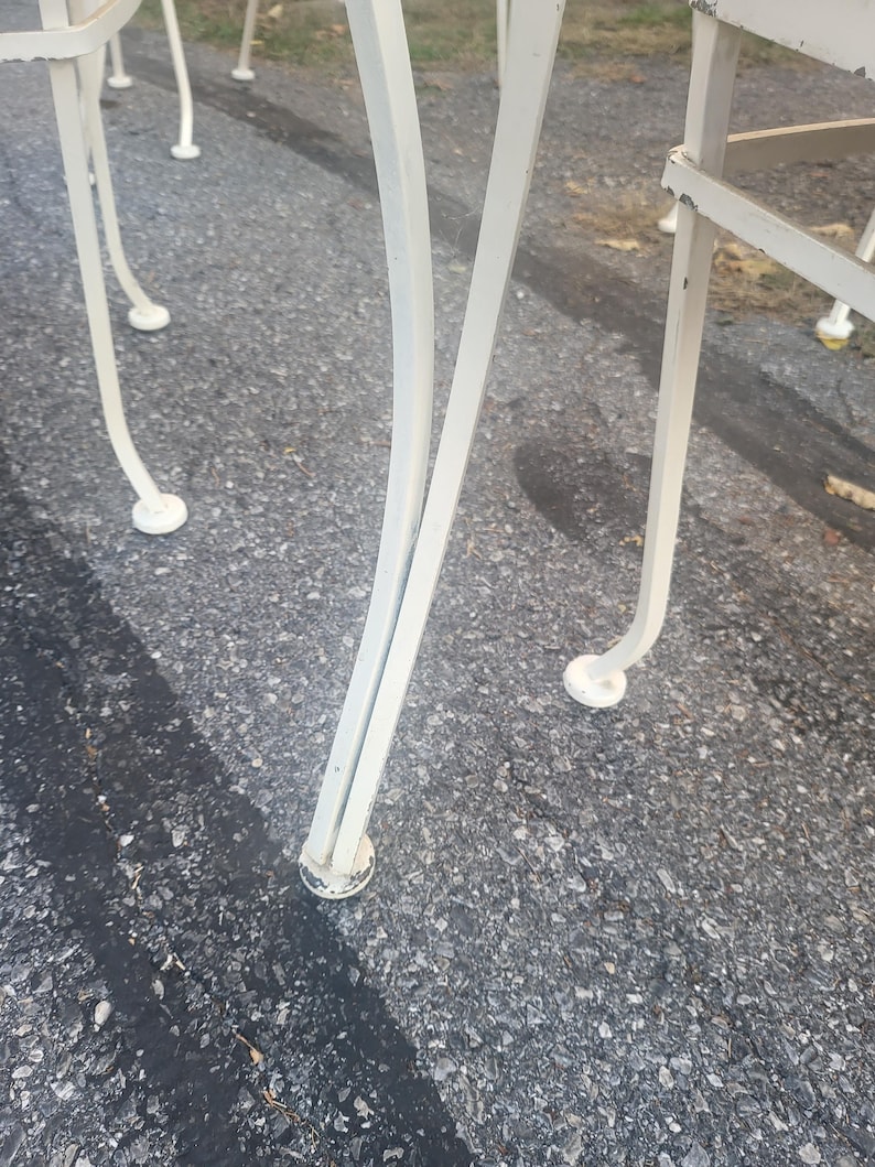 May include: Close-up of the white metal legs of a patio chair. The legs are curved and have a decorative design. The chair is sitting on a gray asphalt surface.