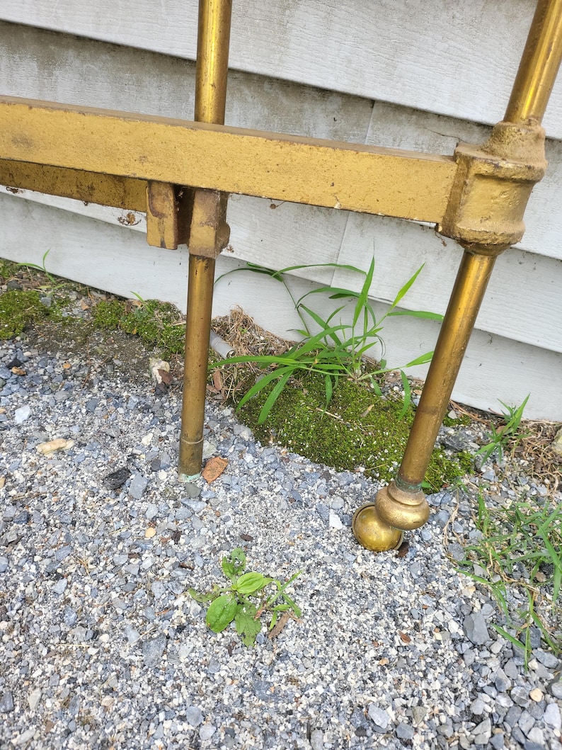 Op de afbeelding: Close-up van een gedeelte van een goudkleurig metalen bedframe. Het frame heeft een rechthoekig hoofdbord en afgeronde poten met kleine zwenkwielen. De poten zijn verbonden met messing fittingen. Het frame staat buiten op een grindoppervlak.