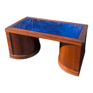 May include: A rectangular wooden coffee table with a dark blue glass top. The table has a rich, warm brown finish and curved legs. The glass top reflects a tree-lined sky, adding a unique visual element to the furniture.