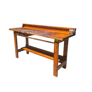 May include: A rectangular wooden workbench with a warm brown finish, featuring a flat work surface, a single drawer on the left side, a raised back ledge, a lower storage shelf, and a small metal pencil sharpener mounted on the right side.