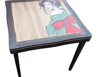 May include: Square wooden side table with a black frame and legs. The tabletop features a painted geisha design with a teal kimono and red bow. The table is approximately 24 inches long and wide, and 20 inches tall.
