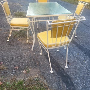 May include: A white metal patio dining set with a glass table top and four chairs. The chairs have yellow cushions.